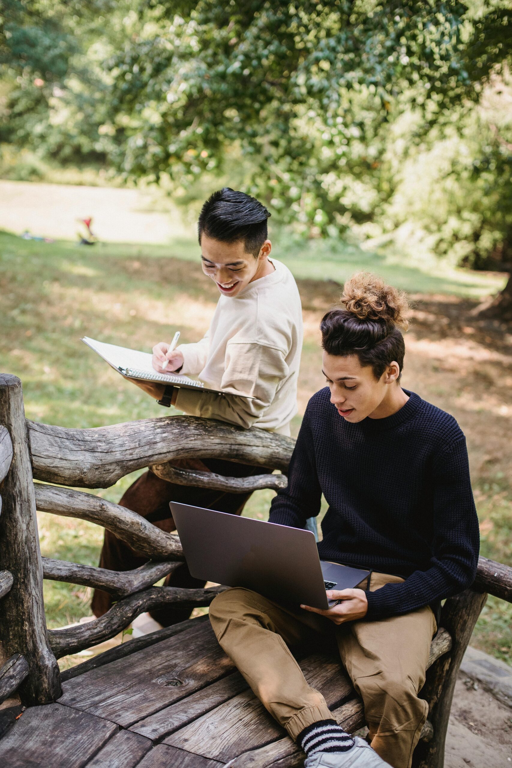 Happy young ethnic male students in casual outfits browsing modern laptop and taking notes in notebook while studying together in sunny park